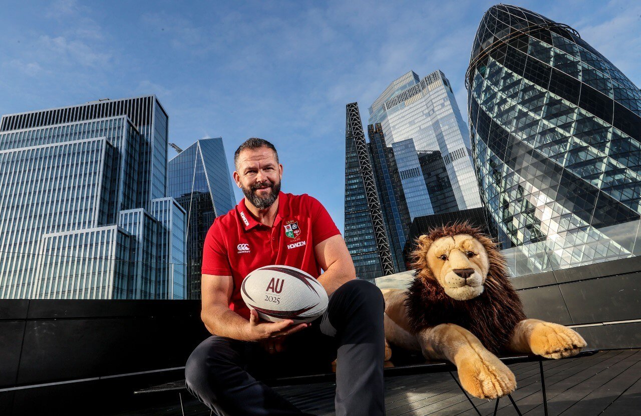 Andy Farrell in a red Lions jersey holds a rugby ball printed with AU 2025, while sitting next to a lion plush toy. Skyscrapers and the Gherkin building are in the background under a clear blue sky.