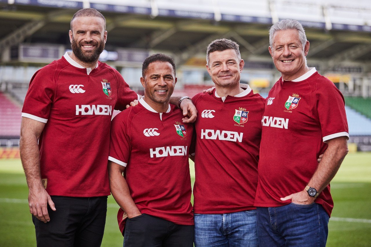 Jamie Roberts, Jason Robinson, Brian O'Driscoll, and Gavin Hastings standing with their arms around each other and smiling in a rugby stadium, with the green rugby pitch showing behind them